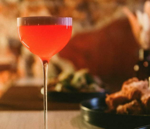 Rød cocktail i elegant glass, med slørete bakgrunn av mat og mørke omgivelser.